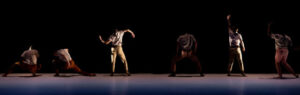 Two BDT dancers facing the back. One, more lit, bends arms forming a wave-like shape.