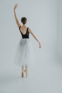Ballet dancer on pointe, in a black leotard and a long white tutu faces the back and lifts one arm up while elongating the other down.