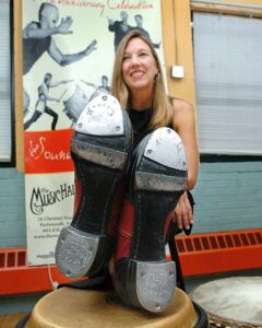 Photo of Drika smiling and crossing legs over a high surface showcasing soles of tap shoes.