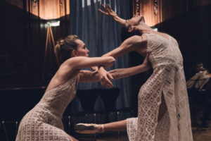 Two dancers in dresses interlock arms while one arches back and the other presses other hand into their back.