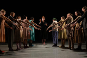 Dancers standing in two diagonal lines forming a V shape extend their arms towards the center while one dancer stands in center with arms elevated as if conducting an orchestra.