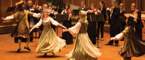 Dancers in baroque costumes twirl around a space with musicians performing.