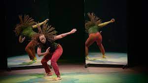 Black female-presenting dancer, her hair flying, stoops as she dances in front of two mirrors that reflect her back.