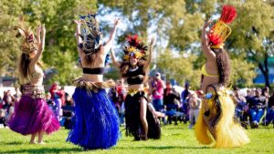 Four Polynesian dancers in colorful raffia skirts raise their arms as they dance outdoors