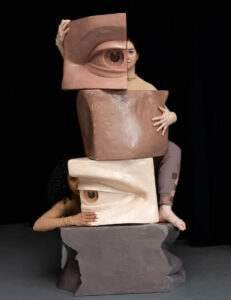 Two dancers interact and hide most of their bodies behind a totem constructed of different cubes with different parts of the human face in different skin tones.