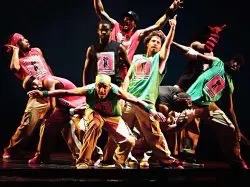 Photo of dancers on stage with loose tank tops and baggy pants gathered closely and reaching arms in different directions.