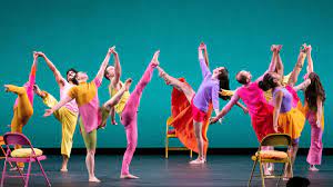 Colorfully dressed Mark Morris dancers, against an intense turquoise backdrop, swing their legs and arms up joyfully.