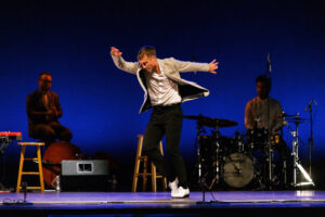 Tap dancer performs in black pants, white button down shirt and open suit jacket while two musicians sit in the background and accompany.