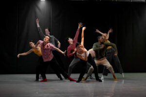 Cirio Collective dancers in a dark space moving as a group reaching arms in different directions.