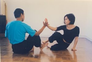 Two dancers sit on the floor looking at each other and press wrists against each other's.