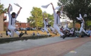 Large group of dancers in all white leap with one leg forward and one leg back.
