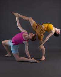 Two dancers: one standing on one leg wearing yellow in a back bend, while the other kneels on one knee and reaches for standing dancer's lifted ankle while also in a back bend.