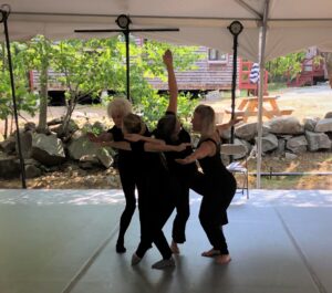 A group of four female dancers dresses in black create a leaning group form with arms outstretched on a grey floored, white canopied pavilion with natural setting in the background.