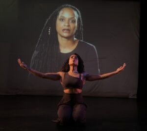 A Large movie screen picture of a Amanda Shea (a poet ) and Dancer on Mayra Hernandez, below her, on her knees arms open wide  looking up.