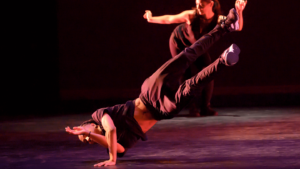 Hip Hop dancer on stage landing an intricate inversion on one shoulder while legs are in the air on a diagonal.