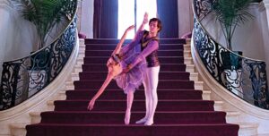 Pas de Deux on stair case: one dancer on pointe and in a purple tutu hinges forward while listing one leg straight behind as the other dancer in white tights and a maroon long sleeve shirt hold on to them from their waist.