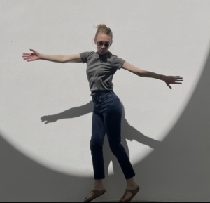 Ana Harmon in a bun, sunglasses, grey t-shirt, jeans and sandals jumps just slightly off the ground into a spotlight and reaches arms wide to the sides as she twists her lower body.