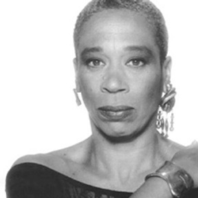 Black and white photo of Adrienne Hawkins wearing large asymmetrical earrings and with one arm across chest displaying large bracelet.