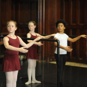 Three children at the barre standing in first position looking over their right shoulders.