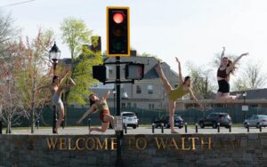 Four dancers pose by "Welcome to Waltham" sign. Traffic light separates them in 2 and 2.