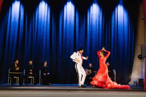 Two flamenco dancers on stage dancing while musicians sit and play their instruments.
