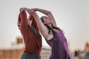Two dancers: one brings hands to shoulders with elbows up while the other, in front, reaches back to touch other dancers elbows.