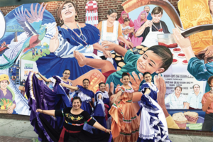 Dancers in typical Salvadorian dresses pose in front of a painted mural with latinx cultural icons.