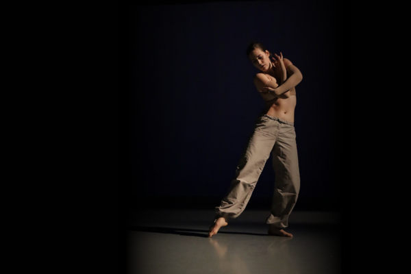 Dancer in a dark space brings arms across body in a self-embrace while extending one leg out to the side.