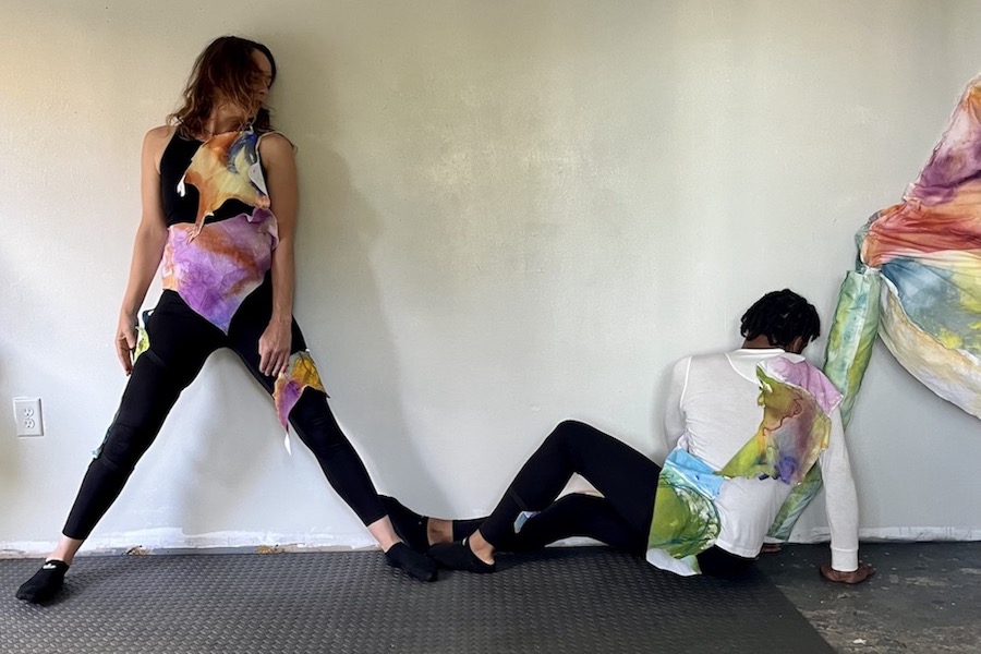 Nora, a white female dancer, stands facing forward while looking towards MacKenzie, a black male dancer, who is sitting beside her with his leg outstretched facing the wall. Their toes connect and they both wear black with fragments of pastel fabric from the soft table like sculpture that is pictured next to MacKenzie.