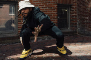A man in a white bucket hat and black tracksuit squats in a dance move on a brick sidewalk in front of a brick wall