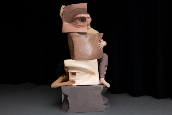 Two dancers behind a totem of sculptures of parts of a human face in different skin tones.