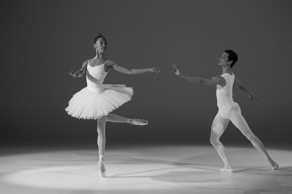 Black and white photo of pas de deux in which male-presenting dancer lunges and reaches towards female-presenting dancer, who is on pointe in aback attitude.