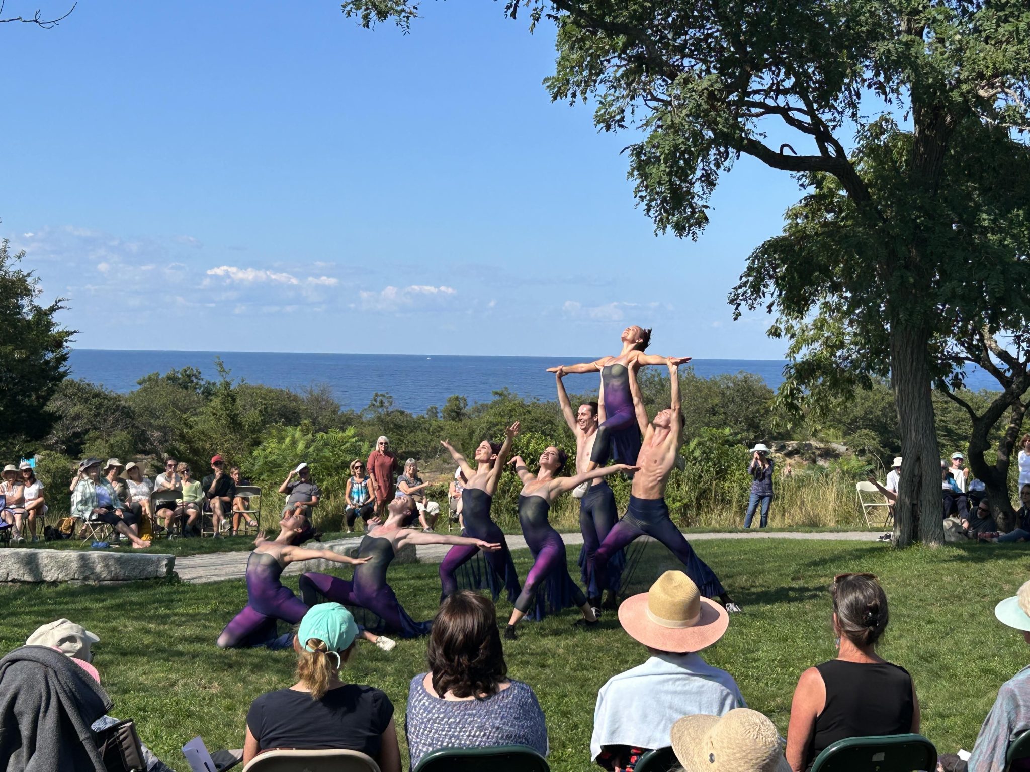 A crowd of people in lawn chairs watch as dancers in body-tight suits tilt their bodies towards the sky; one dancer is held up by two others