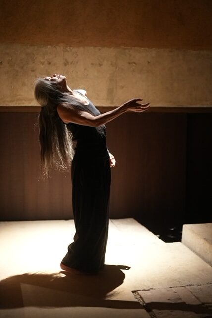 Dancer in dimly lit room looking up to the light with one arm outstretched, palm up.