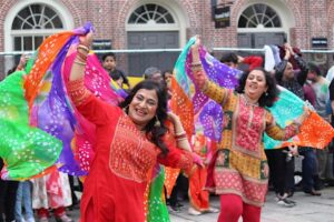 Two dancers move joyfully in bright embroidered outfits, lifting colorful scarves overhead in rhythmic, sweeping gestures. Their bodies twist and extend with expressive energy, surrounded by a lively crowd sharing in the festive dance.