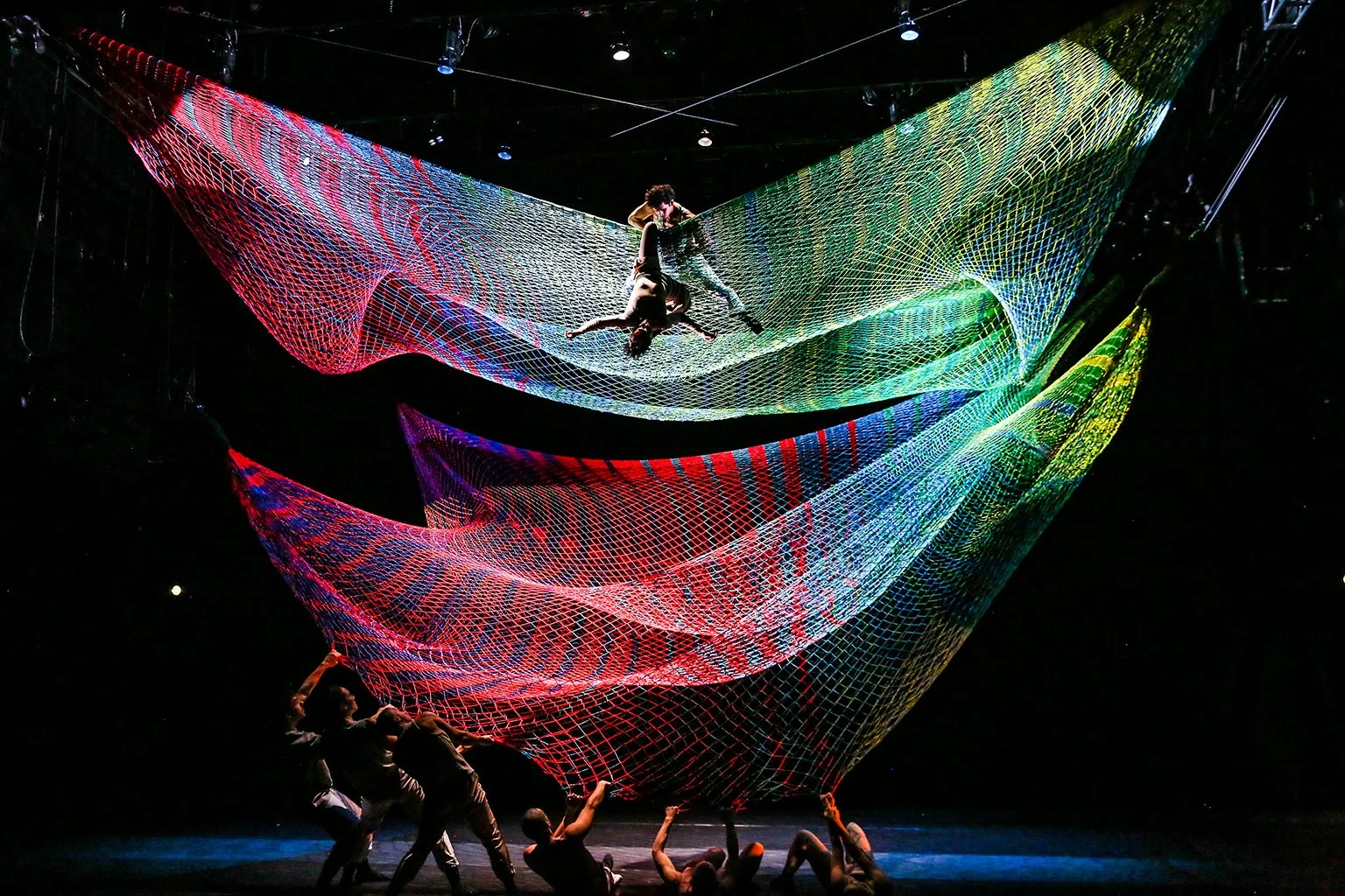 A group of performers engages in an aerial dance performance beneath large, multicolored net structures suspended above the stage. Two dancers balance and intertwine high above the ground on the netting, while others on the stage below hold and manipulate the fabric. The nets are illuminated with vibrant, shifting lights in shades of red, purple, blue, green, and yellow, creating a dynamic, wave-like effect against the dark background.