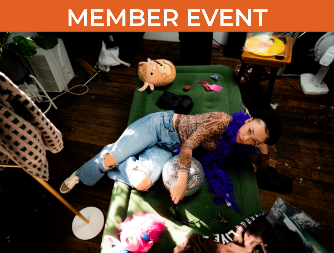 Woman in animal print shirt, blue feather boa, and blue jeans lies on green inflatable mattress with miscellaneous items strewn around