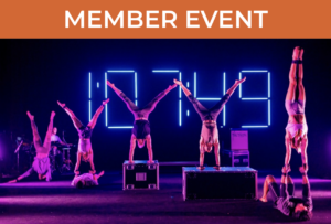 Many artists on stage in handstands, some elevated on boxes and some elevated on other people's bodies. Neon lights in the background spell out "1:07:49" and a set of instruments with someone seated at a keyboard can be seen in the corner.