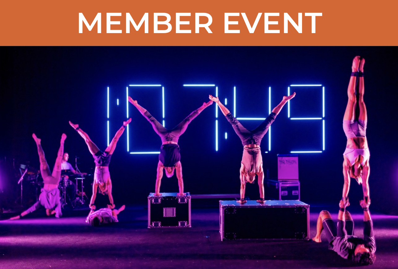 Many artists on stage in handstands, some elevated on boxes and some elevated on other people's bodies. Neon lights in the background spell out "1:07:49" and a set of instruments with someone seated at a keyboard can be seen in the corner.