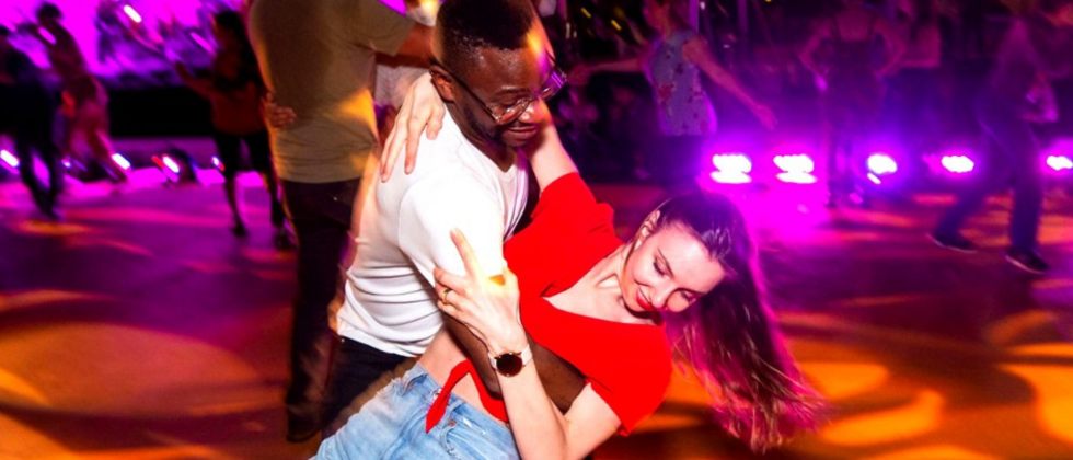 Slightly blurred image of two people dancing joyously together, one dipping the other. Colors are vibrant warm tones with others dancing out of focus in the background.