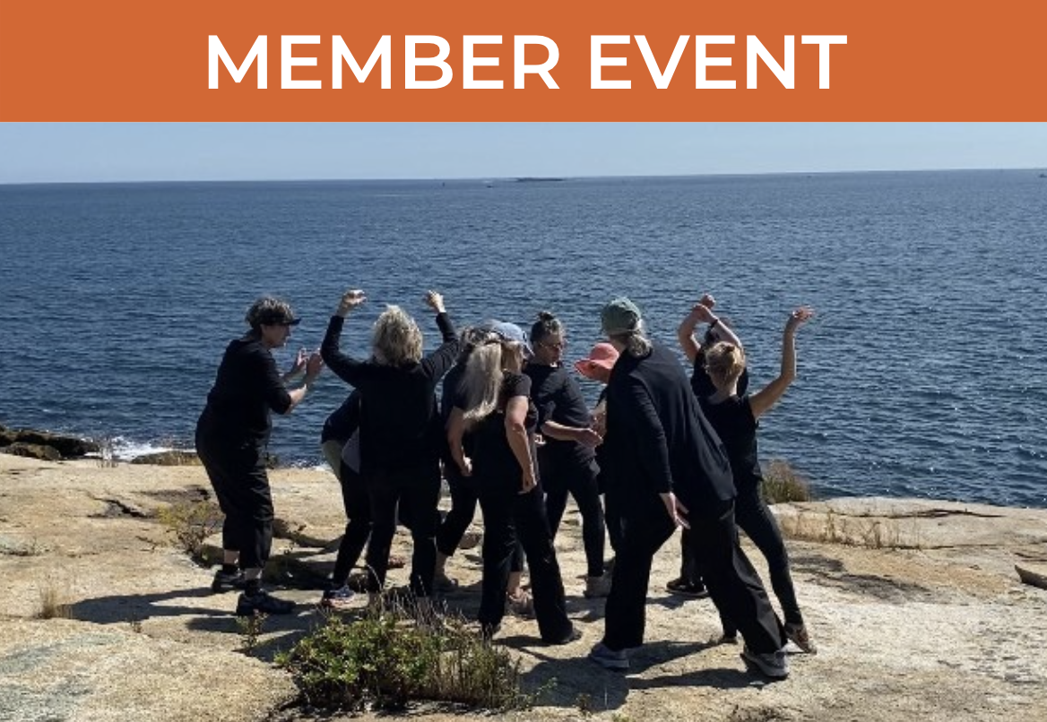 Dancers dressed in black cluster on a rocky coastline with the ocean and sky in the background