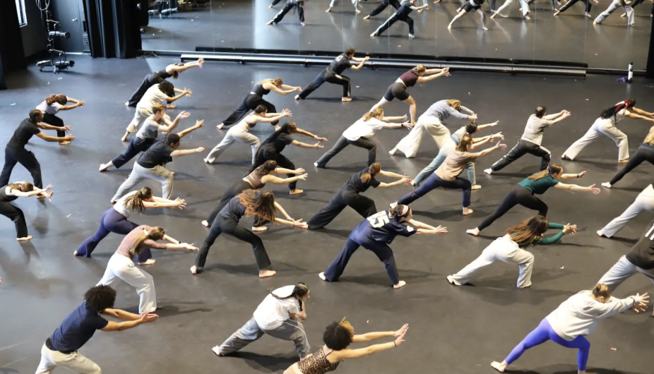 Image taken from above of 20+ dancers all facing the mirror, lunging with arms extended to the side.