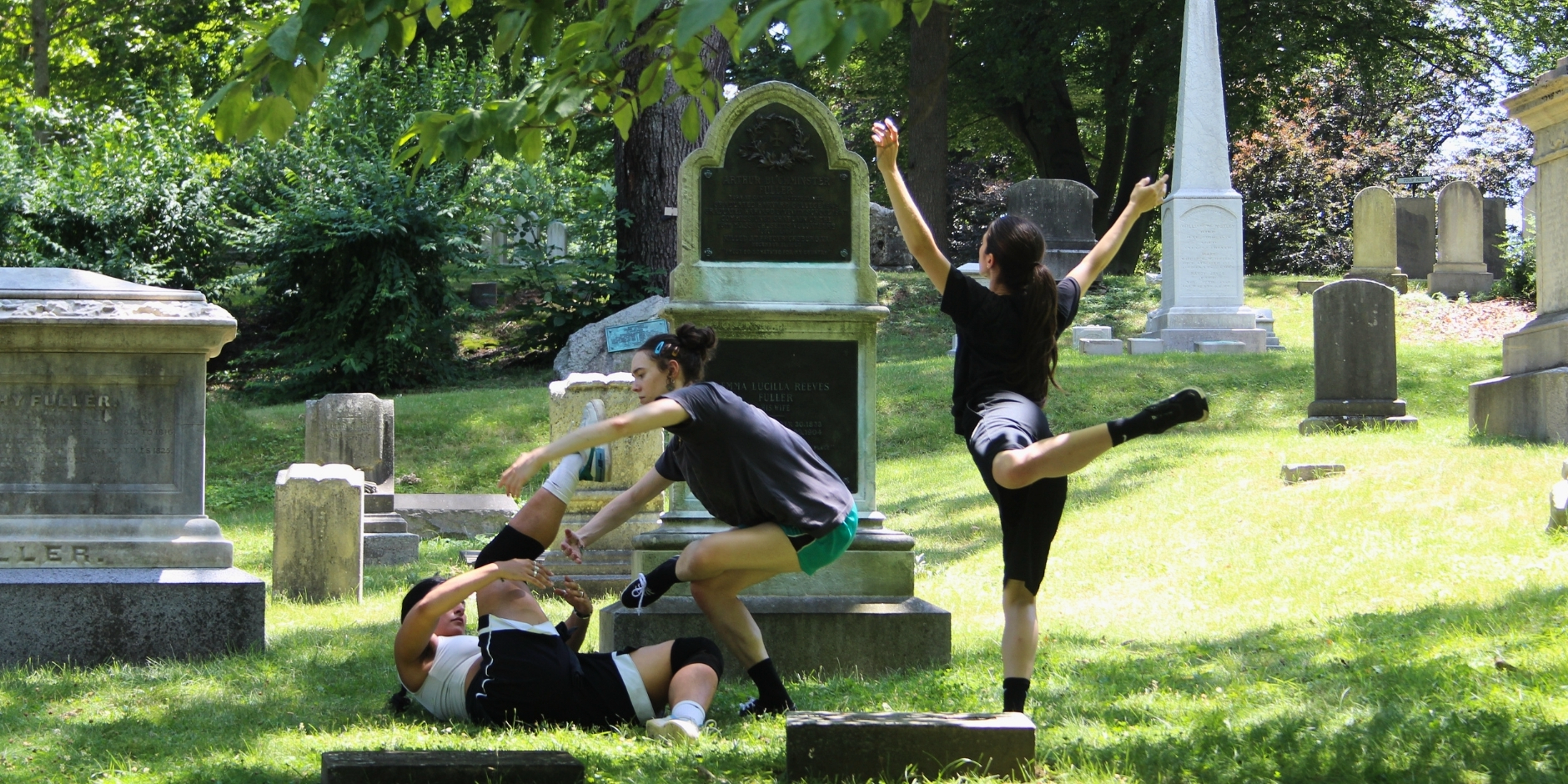 Three dancers in a cemetery dancing on the grass. One dancer is laying on their back with one leg extended to the sky. The seocnd dancer is balancing on one leg and is reaching toward the dancer on the ground. The last dancer looks toward them with their arms extended to the sky, one leg behind them in an attitude, and back to the camera.